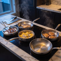 De vijf titanium pannen op een kookplaat, met gebakken vis, worstjes onder een glazen deksel, pasta en kip