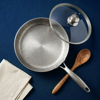Titanium pan met hoge rand van bovenaf, met glazen deksel, houten lepel en linnen servet op een blauwe ondergrond