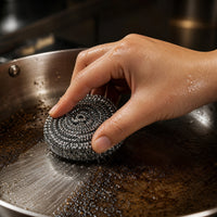 Hand schrobt aangekoekte resten uit een pan met een titanium pannenspons