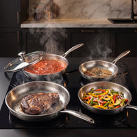 Vier titanium koekenpannen op een kookplaat, met tomatensaus, specerijen, een steak en reepjes paprika