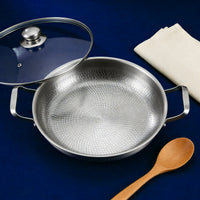 Titanium pan met twee handgrepen van bovenaf, met glazen deksel, houten lepel en servet op een blauwe ondergrond