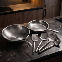 Titanium wok, koekenpan en keukengerei op een donker marmeren aanrecht in een keuken