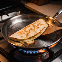 Dubbelgevouwen quesadilla met gesmolten kaas wordt met een houten spatel gekeerd in de titanium crêpepan boven een gasvlam