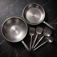 Titanium wok, koekenpan en keukengerei van bovenaf gezien op donker marmer